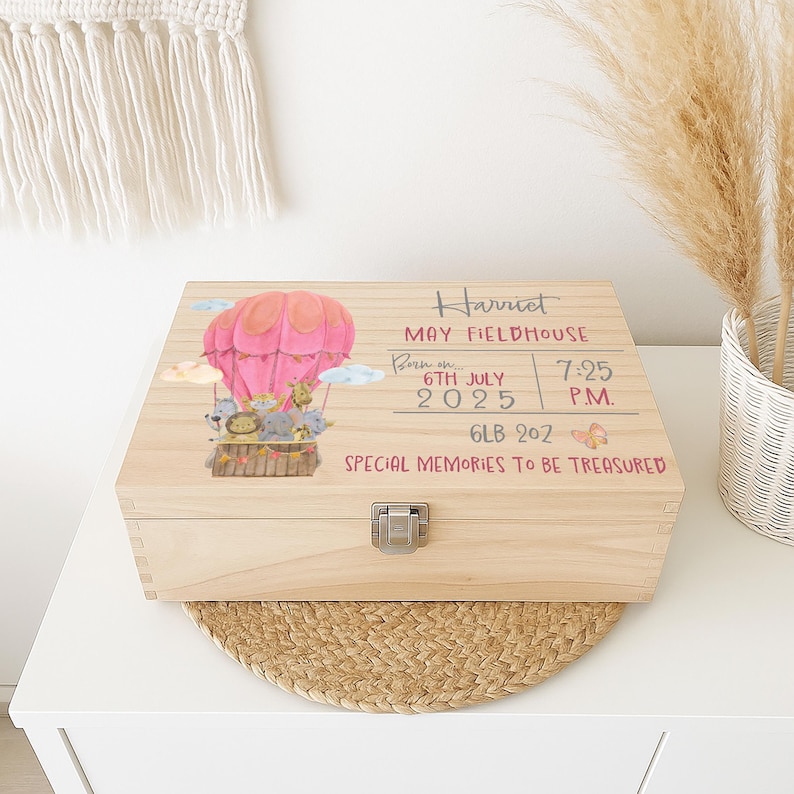 May include: A rectangular wooden keepsake box with a metal latch. The lid features a pink hot air balloon with animal illustrations, and printed text: Harriet, May Fieldhouse, Born on 6th July 2025, 7:25 P.M., 6lb 2oz, Special Memories To Be Treasured.