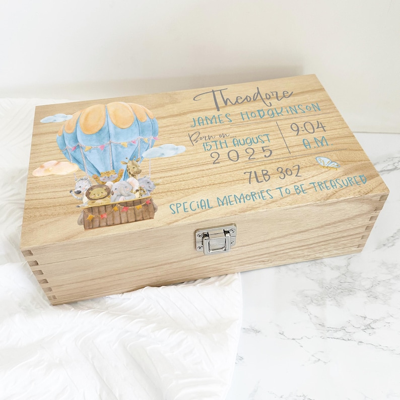 May include: A rectangular wooden keepsake box with a metal latch, featuring a watercolor illustration of a hot air balloon carrying animals. Text includes the name Theodore James Hodgkinson, birth date 15th August 2025, time 9:04 AM, weight 7LB 3OZ, and the phrase SPECIAL MEMORIES TO BE TREASURED.