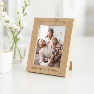 May include: A light-coloured wooden picture frame with the words "WORLD'S BEST Daddy" engraved at the top and "Love Mabel & Oliver" at the bottom. The frame holds a photo of a man and two children.