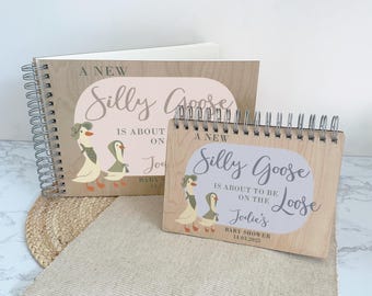 Op de afbeelding: Twee houten gastenboeken met een ganzenmotief. Het grotere boek heeft de tekst "A New Silly Goose is About On" en het kleinere boek heeft de tekst "A New Silly Goose is About To Be On The Loose Jodie's Baby Shower 14.03.2025".