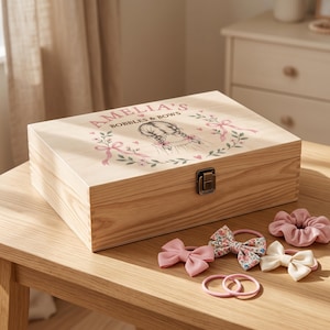 May include: A rectangular light-coloured wooden storage box with a metal latch. The lid features a printed illustration of a person with braided hair, pink ribbons, and green leaves, with the text AMELIA'S BOBBLES & BOWS. Hair accessories are nearby.