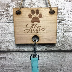 Pu&ograve; includere: Uncino in legno per guinzaglio per cani con un'impronta di zampa e il nome "Alfie" inciso. L'uncino &egrave; attaccato a un guinzaglio blu chiaro.