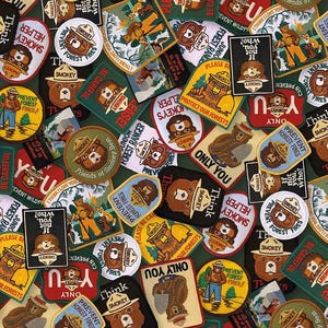 May include: A collection of Smokey Bear patches in various shapes and sizes. The patches feature Smokey Bear in a ranger hat, along with text like "Prevent Forest Fires" and "Only You." The color palette includes brown, yellow, red, and green.