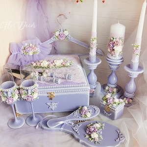 Puede incluir: Un conjunto de boda lavanda con una caja, dos copas de champán, una percha de madera, dos velas, una caja pequeña y un cepillo para el cabello. El conjunto está decorado con encaje, perlas y flores.