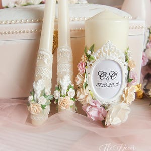 Puede incluir: Dos velas de boda blancas decoradas con encaje y detalles florales. La vela más grande tiene un marco blanco con las iniciales "O & O" y la fecha "27.10.2022".