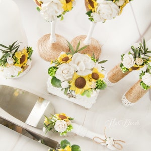 May include: A wedding cake cutting set with a white knife handle and a silver blade. The handles of the knife and server are decorated with white flowers, green leaves, and yellow sunflowers. The set is displayed on a white surface with a small white box decorated with flowers and a burlap ribbon.