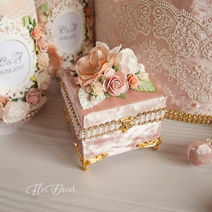 Könnte beinhalten: Eine rosafarbene Samt-Ringbox mit goldener Verzierung und Perlenakzenten. Die Box ist mit einem Strauß aus pfirsichfarbenen, weißen und rosafarbenen Stoffblumen verziert.