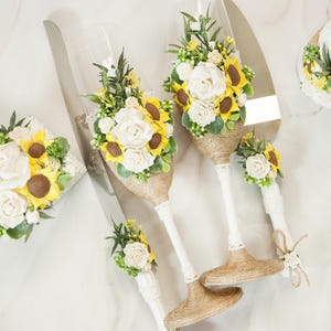 May include: A set of two white champagne flutes with burlap ribbon and sunflower and white rose floral accents. A matching cake knife with a similar floral design is also included.