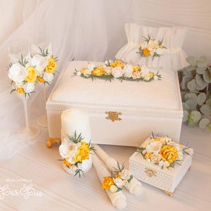 Op de afbeelding: Een verzameling witte huwelijkaccessoires, waaronder een grote doos, een kleine doos, twee kaarsen en twee champagneglazen, allemaal versierd met gele en witte bloemstukken. Op de achtergrond is een witte sluier te zien.