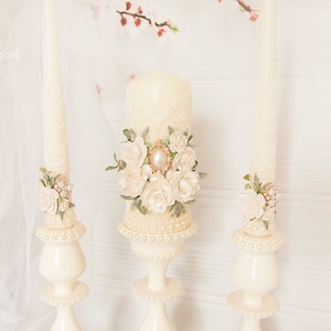 May include: Three ivory wedding candles with pearl accents and floral embellishments. The center candle is taller than the two flanking candles.