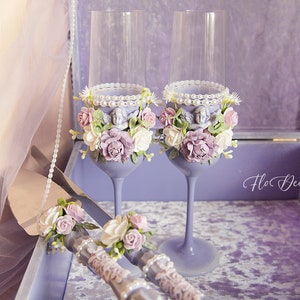 Könnte beinhalten: Zwei lavendelfarbene Champagnerflöten, verziert mit weißen und violetten Blumen, Perlen und kleinen Engelsfiguren. Ein silbernes Kuchenschaber mit passendem Blumendesign ist ebenfalls enthalten. Das Set wird in einer mit violettem Samt ausgekleideten Schachtel präsentiert.