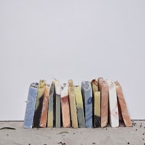 May include: A collection of handmade soap bars in various colors and patterns. The soaps are arranged in a row, leaning against a white background. Colors include blue, black, pink, and yellow, with visible textures and embedded seeds.