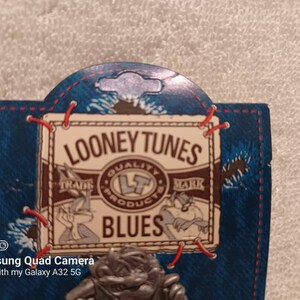 May include: A Looney Tunes pin featuring Bugs Bunny and Daffy Duck. The pin is attached to a blue denim-style backing with red stitching and the words "Looney Tunes Blues" printed on it. The pin has a silver finish.