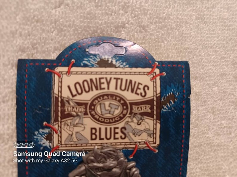 May include: A Looney Tunes pin featuring Bugs Bunny and Daffy Duck. The pin is attached to a blue denim-style backing with red stitching and the words "Looney Tunes Blues" printed on it. The pin has a silver finish.