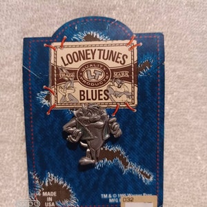 May include: A silver-colored Taz pin from Looney Tunes, attached to a blue denim-style backing. The backing features the Looney Tunes logo and the word "BLUES". The pin is a collectible item, with the text "MADE IN USA" and copyright information.