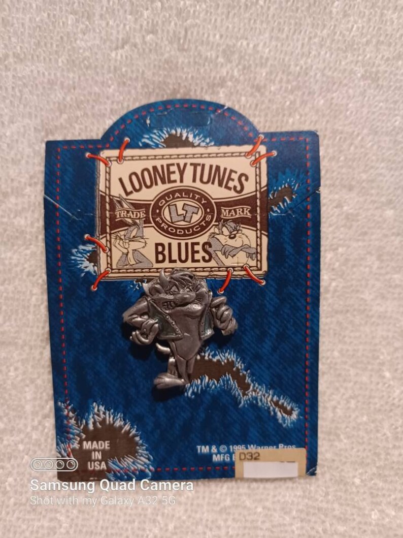 May include: A silver-colored Taz pin from Looney Tunes, attached to a blue denim-style backing. The backing features the Looney Tunes logo and the word "BLUES". The pin is a collectible item, with the text "MADE IN USA" and copyright information.
