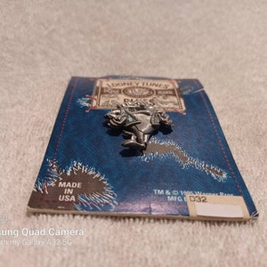 May include: A silver-colored Looney Tunes character pin, possibly a Bugs Bunny character, is displayed on a blue card with the text "LOONEY TUNES" and "MADE IN USA". The pin is a vintage collectible.