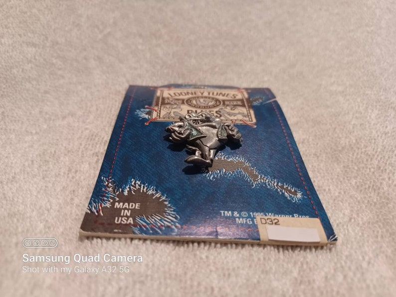May include: A silver-colored Looney Tunes character pin, possibly a Bugs Bunny character, is displayed on a blue card with the text "LOONEY TUNES" and "MADE IN USA". The pin is a vintage collectible.
