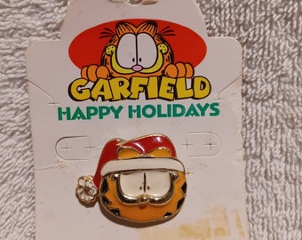 May include: A Garfield pin featuring the cartoon cat wearing a Santa hat, set against a white card with the words "GARFIELD HAPPY HOLIDAYS." The pin is orange, white, and red. The card also includes the text "MFG. IN THE USA BY STARLINE."
