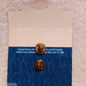May include: Two gold-colored pin backs attached to a blue and white patterned card. The card has text that reads "Looney Tunes" and "Warner Bros. Inc. &copy; 1995". The card is white on the top and has a hole for hanging.