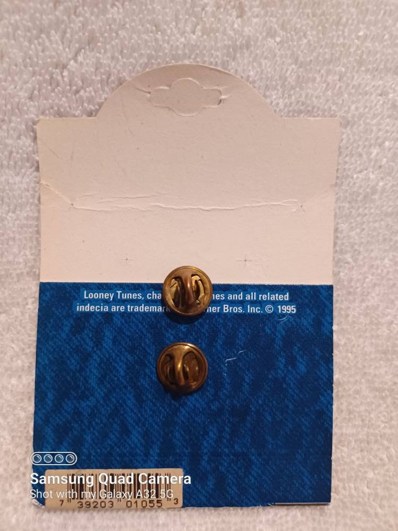 May include: Two gold-colored pin backs attached to a blue and white patterned card. The card has text that reads "Looney Tunes" and "Warner Bros. Inc. &copy; 1995". The card is white on the top and has a hole for hanging.