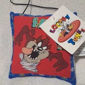 May include: A square, red Taz-Mania Looney Tunes pillow with a blue border. The pillow features a cartoon image of the Tasmanian Devil in a tornado. A white tag with Looney Tunes characters is attached.