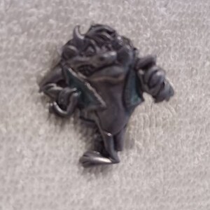 May include: A small, pewter-colored figurine of a horned creature holding a blade. The figure has a hunched posture and is detailed with textured surfaces. The creature is standing on a white, textured surface.