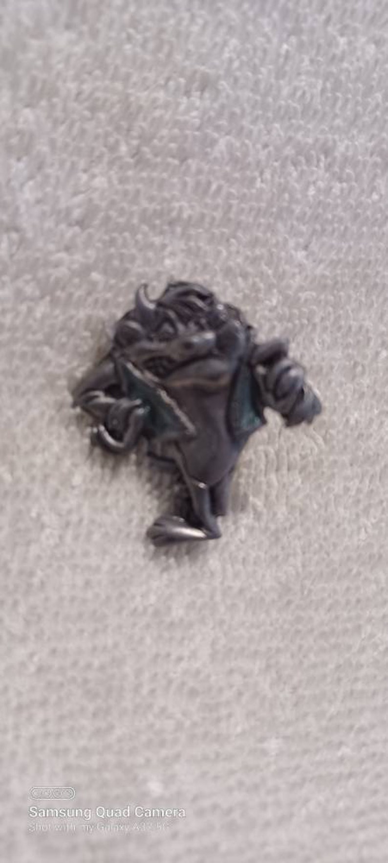 May include: A small, pewter-colored figurine of a horned creature holding a blade. The figure has a hunched posture and is detailed with textured surfaces. The creature is standing on a white, textured surface.