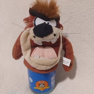 May include: A plush Taz the Tasmanian Devil toy with a brown head, black nose, and open mouth, atop a blue Looney Tunes branded container. The toy has brown fur and long whiskers. The container has the Looney Tunes logo.