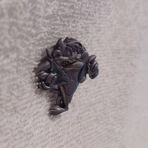 May include: A silver-colored pin featuring the Tasmanian Devil cartoon character. The pin depicts the character in a dynamic pose, with a detailed design and a textured finish. The pin is a collectible item.
