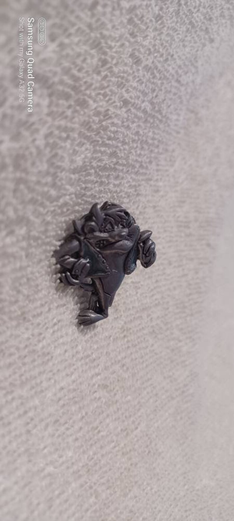 May include: A silver-colored pin featuring the Tasmanian Devil cartoon character. The pin depicts the character in a dynamic pose, with a detailed design and a textured finish. The pin is a collectible item.