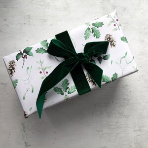 May include: A white gift box with a green velvet bow. The box is wrapped in paper with a pattern of green leaves, berries, and pine cones.