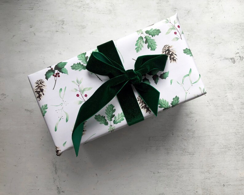 May include: A white gift box with a green velvet bow. The box is wrapped in paper with a pattern of green leaves, berries, and pine cones.