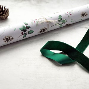 May include: A roll of white wrapping paper with a green and red holly leaf and berry pattern. A green velvet ribbon is lying on a white surface.