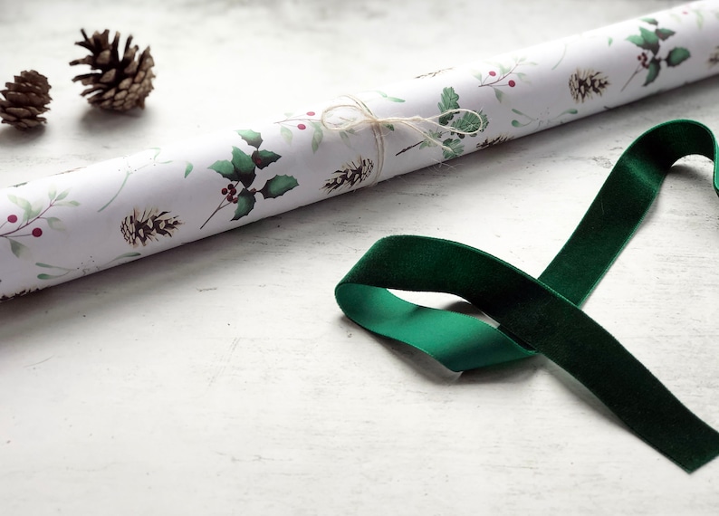 May include: A roll of white wrapping paper with a green and red holly leaf and berry pattern. A green velvet ribbon is lying on a white surface.