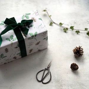 May include: A white gift box with a green ribbon and a green and white floral pattern. The box is sitting on a white surface with two small pine cones and a pair of small scissors.