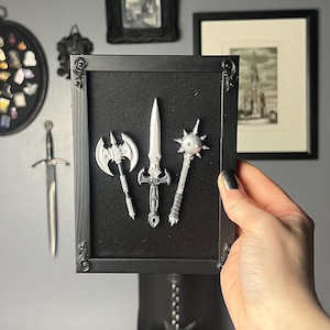 May include: A framed black shadow box featuring miniature silver weapons: an axe, a sword, and a mace. The frame is held against a gray wall adorned with framed art and a Bauhaus poster. The overall aesthetic is gothic and artistic.