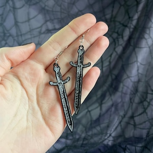 May include: A pair of silver dangle earrings with black sword charms. The swords have a white outline and are detailed with lines.