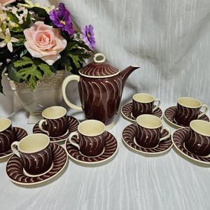 May include: A vintage brown and cream-colored coffee set with a teapot, eight cups, and saucers. The set features a repeating white swirl design. A vase of flowers is in the background.