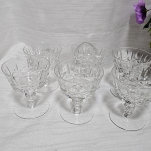 May include: A set of five clear glass coupes featuring a faceted geometric pattern on the bowls and short stems with circular bases. Each glass has a wide, shallow rim and a textured exterior design that catches the light, creating a clear appearance.