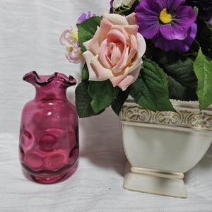 Peut inclure: Un petit vase en verre magenta translucide avec un bord ondulé et un motif circulaire en creux sur le corps, placé à côté d'une jardinière en céramique crème remplie de roses artificielles roses et de pensées violet foncé.