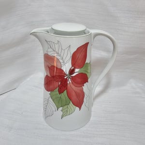 May include: A white ceramic pitcher with a lid and a curved handle, featuring a decorative illustration of red and green poinsettia leaves on the front. The design includes both solid coloured leaves and outlined leaf sketches on the body.