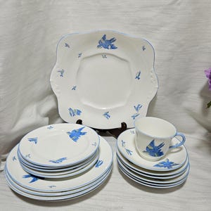 Puede incluir: Un juego de vajilla de cerámica con base blanca, ilustraciones de pájaros azules y borde azul claro. El conjunto incluye una fuente de servir cuadrada grande, varios platos llanos redondos, platos pequeños y una taza con platillo.