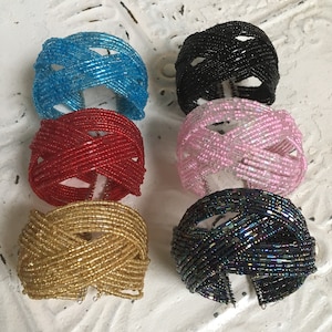 May include: Six beaded bracelets in various colours, including turquoise, red, pink, gold, black, and iridescent black. The bracelets are made of small beads woven together in a criss-cross pattern.