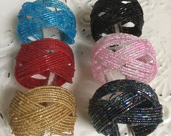 May include: Six beaded bracelets in various colors, including turquoise, red, pink, gold, black, and iridescent black. The bracelets are made of small beads woven together in a criss-cross pattern.