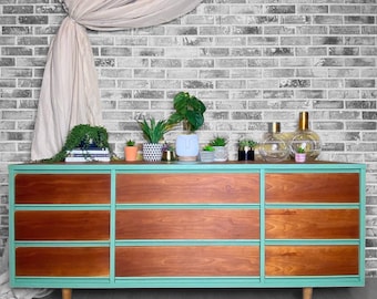 Puede incluir: Una cómoda de seis cajones con un acabado pintado de verde claro y frentes de cajones de madera. La cómoda tiene un estilo moderno de mediados de siglo y está sentada sobre un suelo de madera clara. La cómoda está decorada con plantas y jarrones.