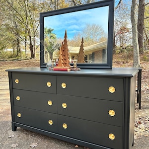 May include: A black dresser with six drawers and gold knobs. The dresser has a mirror on top with a reflection of trees and a blue sky.
