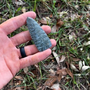 May include: A grey arrowhead with a textured surface, held in a hand. The stone arrowhead has a sharp point. The background is blurred with green grass and brown leaves.