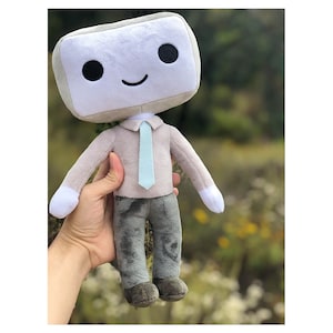 Puede incluir: Un juguete de peluche con una cabeza en forma de pantalla gris y blanca, sonriendo con ojos negros. El juguete lleva una camisa gris con una corbata azul claro y pantalones grises.