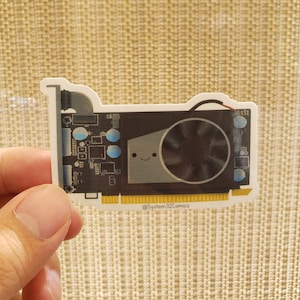 May include: A sticker of a cartoon graphics card with a smiling face and a black fan. The sticker has a yellow border and the text "@System32Comics" at the bottom.